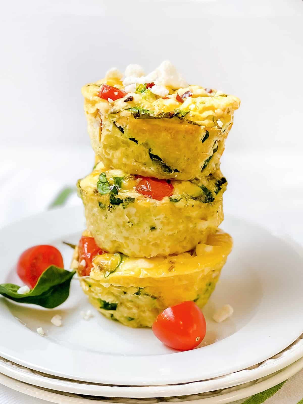 Three delicious egg muffins with spinach, tomatoes, and cheese stacked on a white plate, garnished with cherry tomatoes and fresh spinach leaves.