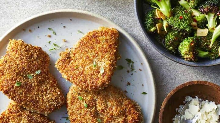 A plate with four breaded chicken cutlets, a bowl of roasted broccoli, a small bowl of grated cheese, and two glasses of white wine—perfect for April Clean Eating Dinners on a gray surface.
