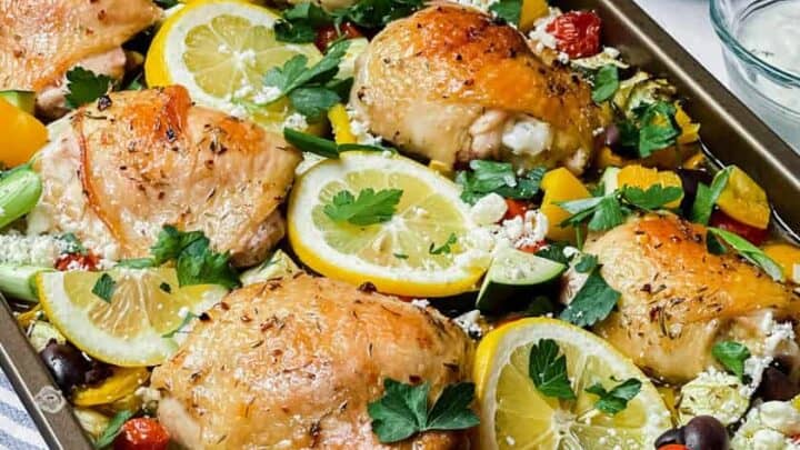 Side view of baked Greek Chicken on a sheet pan with tomatoes and slices of lemon