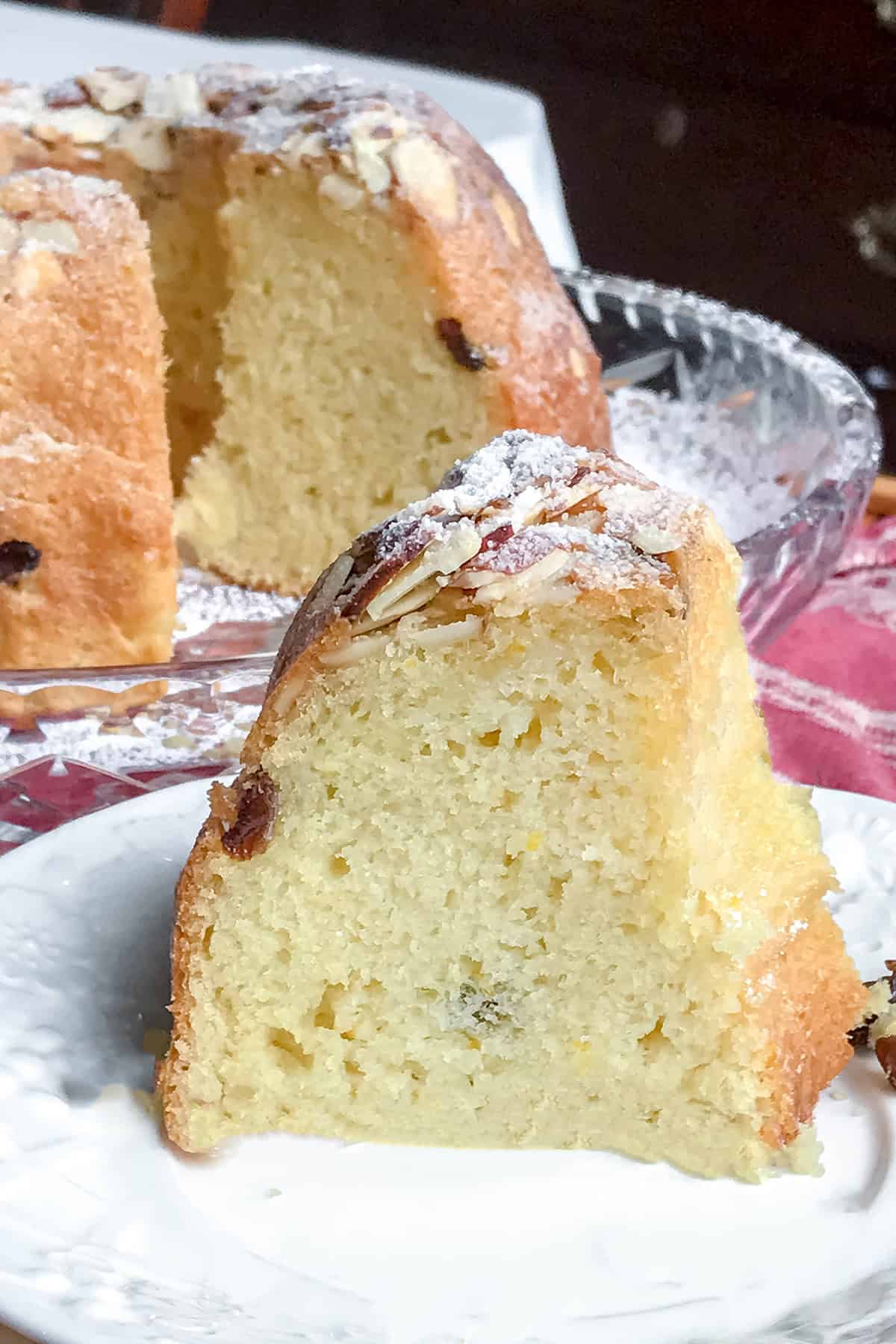 A slice of yellow kugelhopf sponge cake with a light dusting of powdered sugar and almonds, served on a white plate with the rest of the cake in the background.