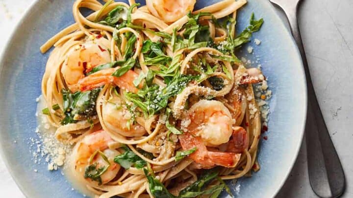 A plate of linguine pasta topped with shrimp, arugula, and grated cheese—one of those perfect 20 Minute Healthy Spring Dinners—served with a fork and knife on the side.