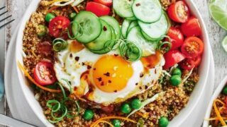 A bowl of quinoa salad topped with a sunny-side-up egg, cucumber, cherry tomatoes, green peas, and scallions—perfect for quick 20 Minute Healthy Spring Dinners. A fork rests beside the bowl.