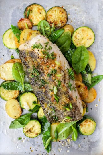 Baked Fish on a sheet pan with vegetables, fresh herbs, and salt and pepper ready for serving.