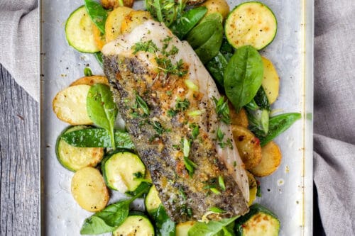 Baked Fish on a sheet pan with vegetables, fresh herbs, and salt and pepper ready for serving.