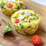 Two egg muffins with spinach, tomato, and feta cheese sit on a wooden board, highlighting the fresh ingredients in these delicious egg muffins. A spinach leaf and a halved cherry tomato rest nearby for a vibrant touch.