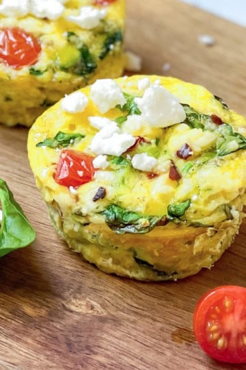 Two egg muffins with spinach, tomato, and feta cheese sit on a wooden board, highlighting the fresh ingredients in these delicious egg muffins. A spinach leaf and a halved cherry tomato rest nearby for a vibrant touch.