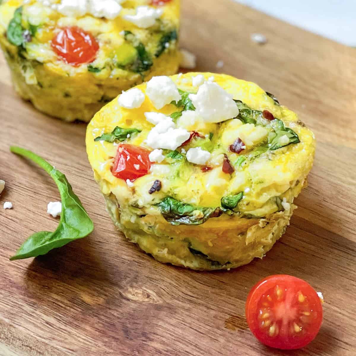 Two egg muffins with spinach, tomato, and feta cheese sit on a wooden board, highlighting the fresh ingredients in these delicious egg muffins. A spinach leaf and a halved cherry tomato rest nearby for a vibrant touch.