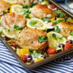 Square image of baked and brown Greek chicken thighs, vegetables, tomatoes and lemon slices on a sheet pan ready for serving.