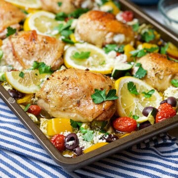 Square image of baked and brown Greek chicken thighs, vegetables, tomatoes and lemon slices on a sheet pan ready for serving.