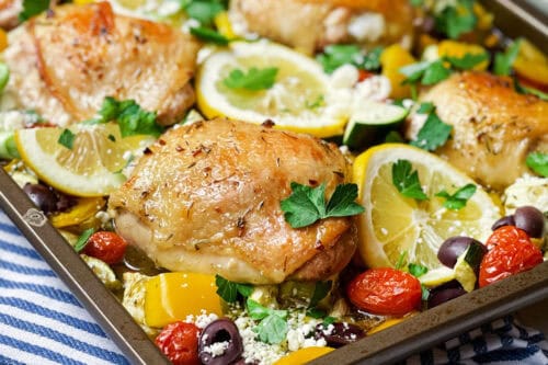 Square image of baked and brown Greek chicken thighs, vegetables, tomatoes and lemon slices on a sheet pan ready for serving.