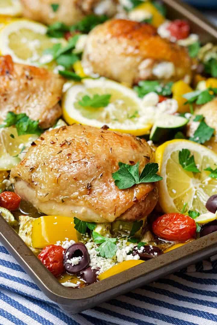 Square image of baked and brown Greek chicken thighs, vegetables, tomatoes and lemon slices on a sheet pan ready for serving.