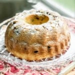 A classic kugelhopf bundt cake, dusted with powdered sugar, sits on a glass platter with a red patterned cloth underneath.