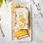A rectangular lemon drizzle loaf cake with white icing and lemon slices on top, presented on a decorative white tray with one slice cut, next to a fork and some white flowers.