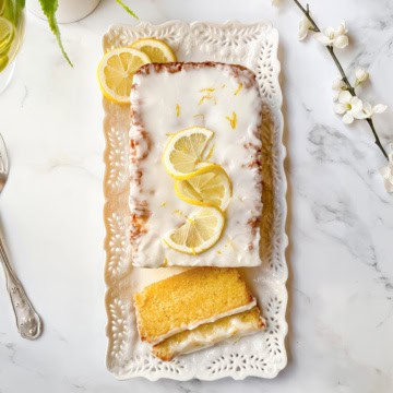 A rectangular lemon drizzle loaf cake with white icing and lemon slices on top, presented on a decorative white tray with one slice cut, next to a fork and some white flowers.
