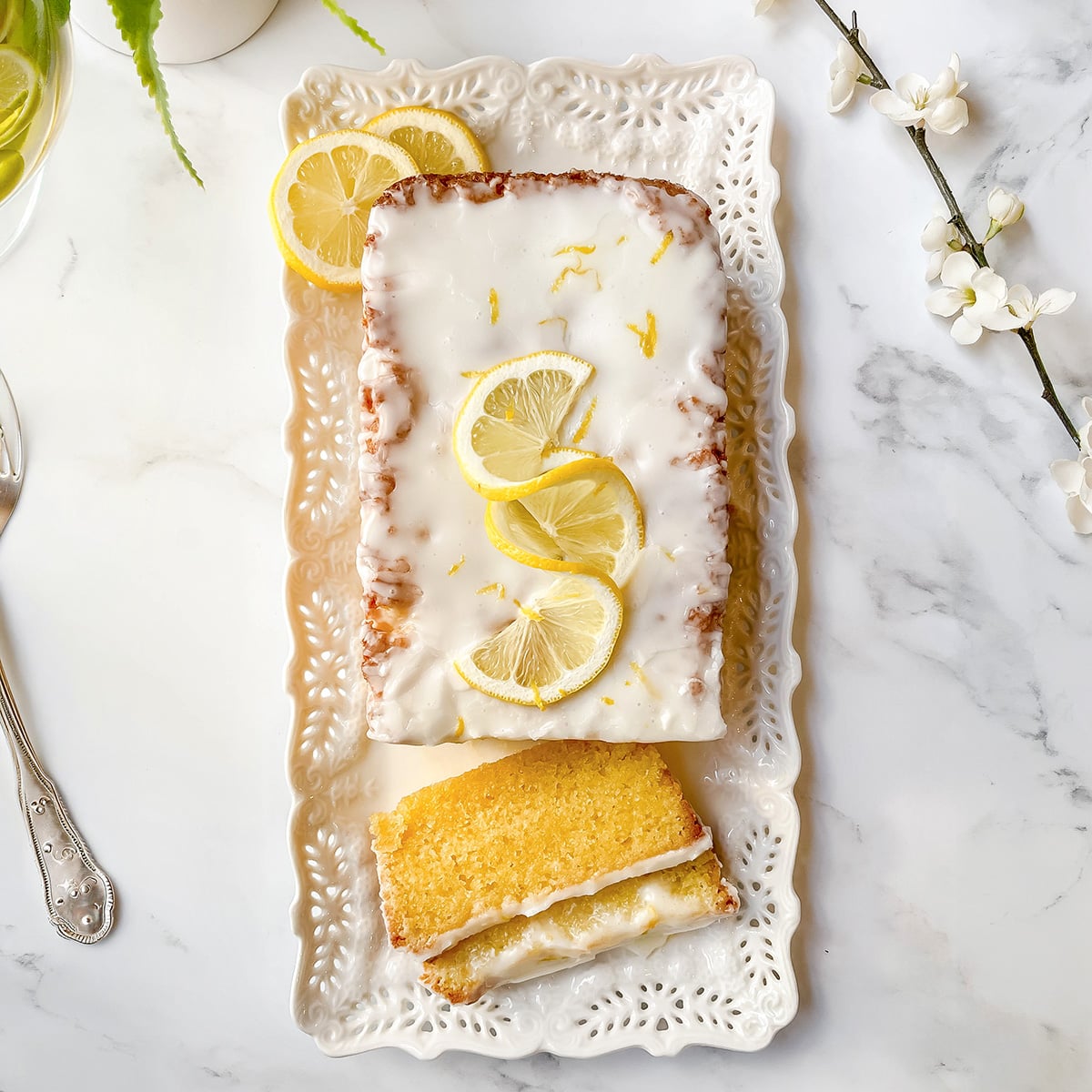 A rectangular lemon drizzle loaf cake with white icing and lemon slices on top, presented on a decorative white tray with one slice cut, next to a fork and some white flowers.