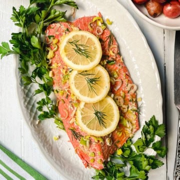 Baked salmon fillet garnished with lemon slices, herbs, and chopped leeks, presented on a white oval platter with fresh parsley around the edges.