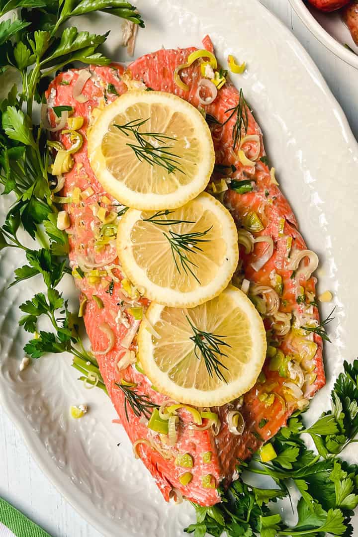 Baked salmon fillet garnished with lemon slices, herbs, and chopped leeks, presented on a white oval platter with fresh parsley around the edges.