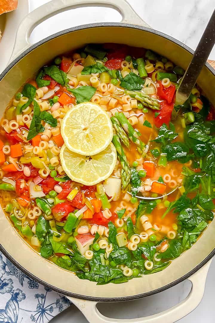A pot of spring minestrone soup with pasta, spinach, tomatoes, carrots, and sliced lemon on top. A ladle rests in the pot, accompanied by fresh bread and herbs on the side.