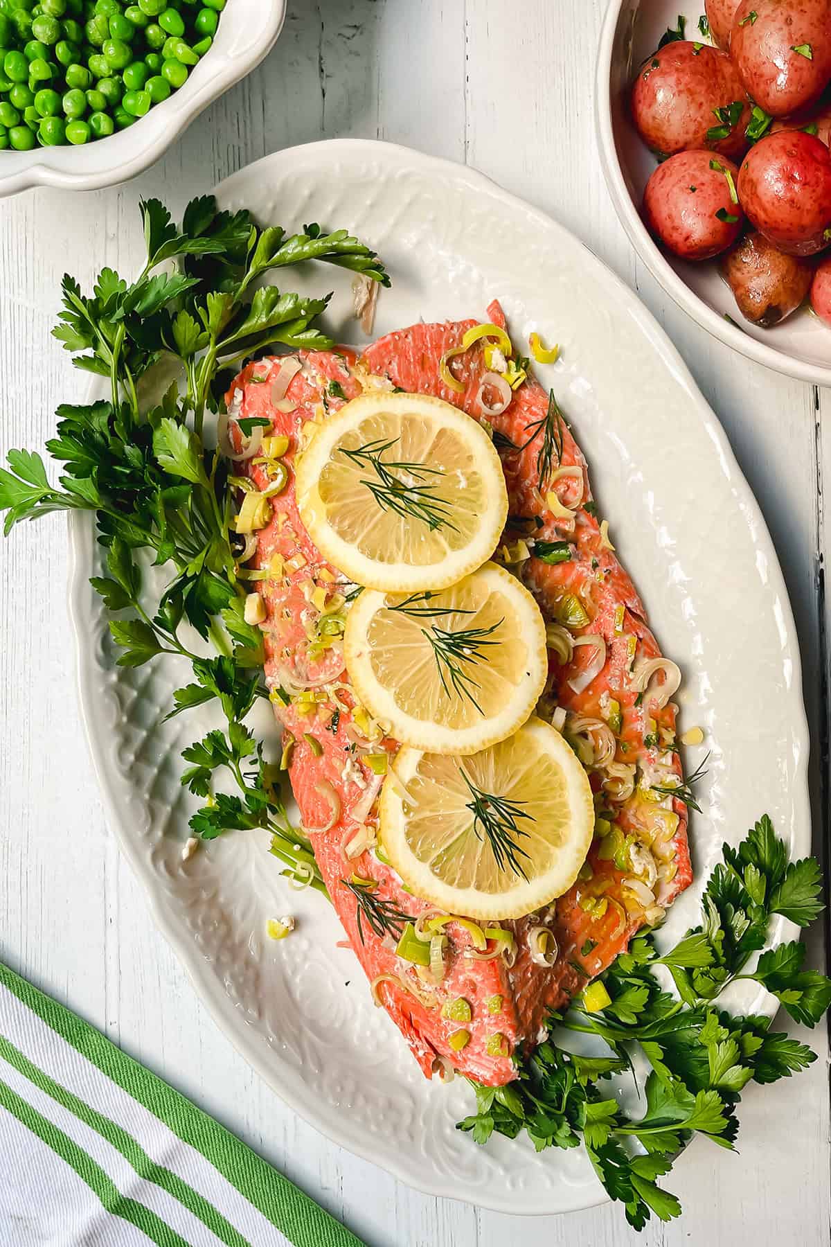 Baked salmon fillet on a white platter, garnished with three lemon slices, dill, chopped leeks, and fresh parsley, with bowls of peas and red potatoes nearby.