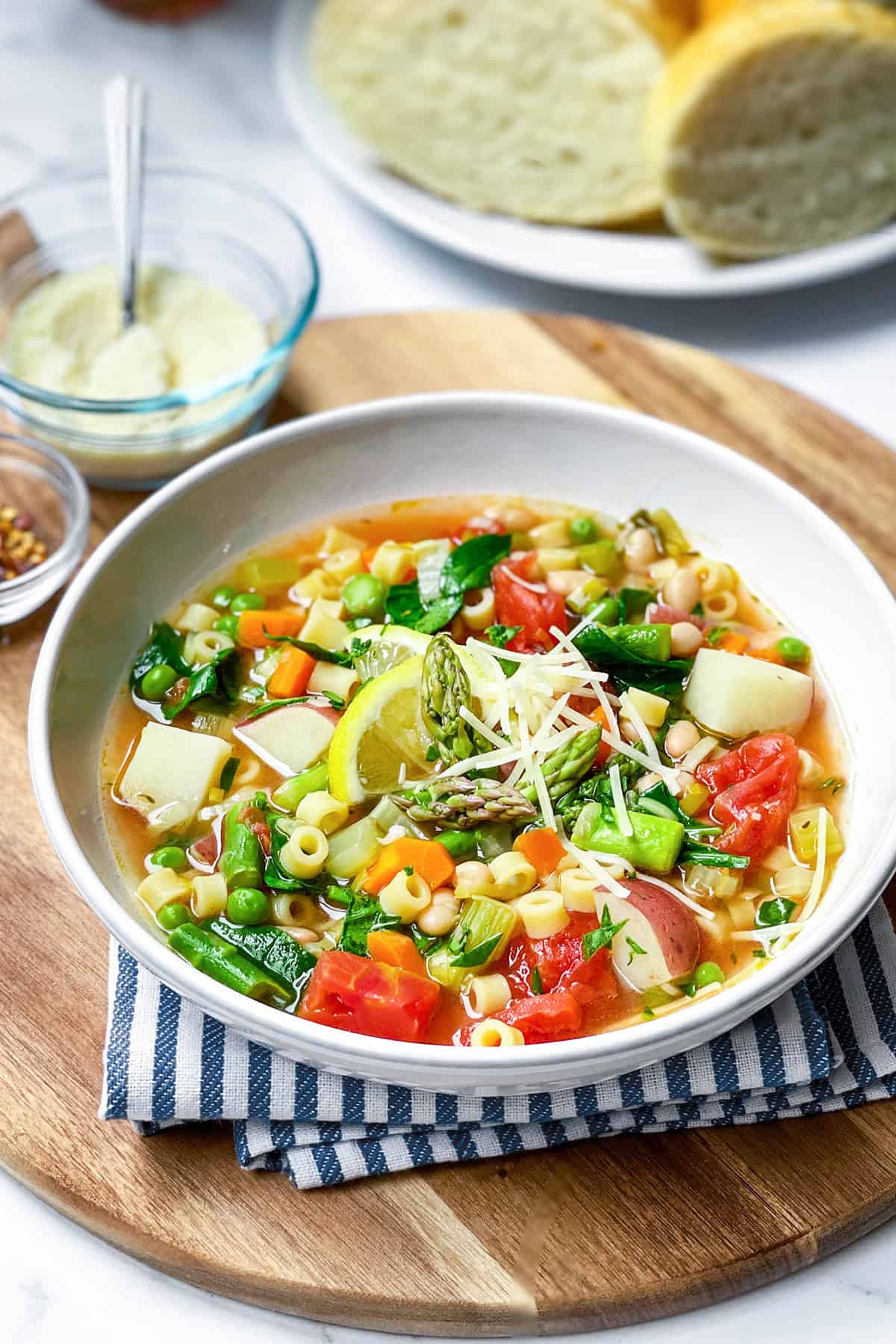 A bowl of spring minestrone soup with pasta, potatoes, tomatoes, and green vegetables, garnished with lemon and grated cheese, on a wooden board with bread slices in the background.
