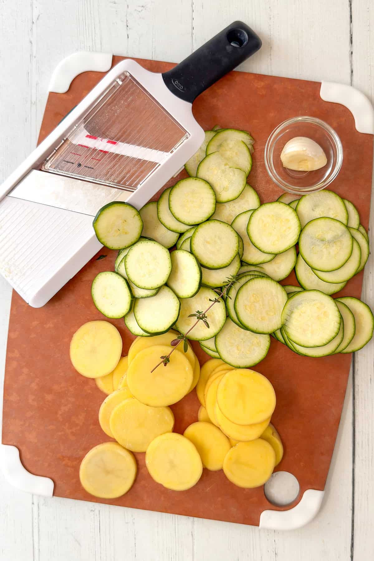 Thinly sliced zucchini and Yukon gold potatoes on a cutting board with a handheld mandolin.