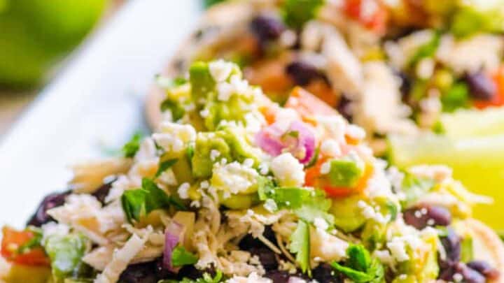 Two tostadas topped with shredded chicken, black beans, avocado, diced red onion, tomato, cilantro, and crumbled cheese—perfect for April Clean Eating Dinners—served on a white plate with lime halves in the background.