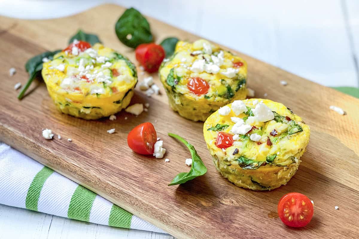 Three egg muffins with spinach, cherry tomatoes, and feta cheese are arranged on a wooden board, with fresh spinach leaves and tomato halves scattered around, making these egg muffins a colorful and appetizing snack.