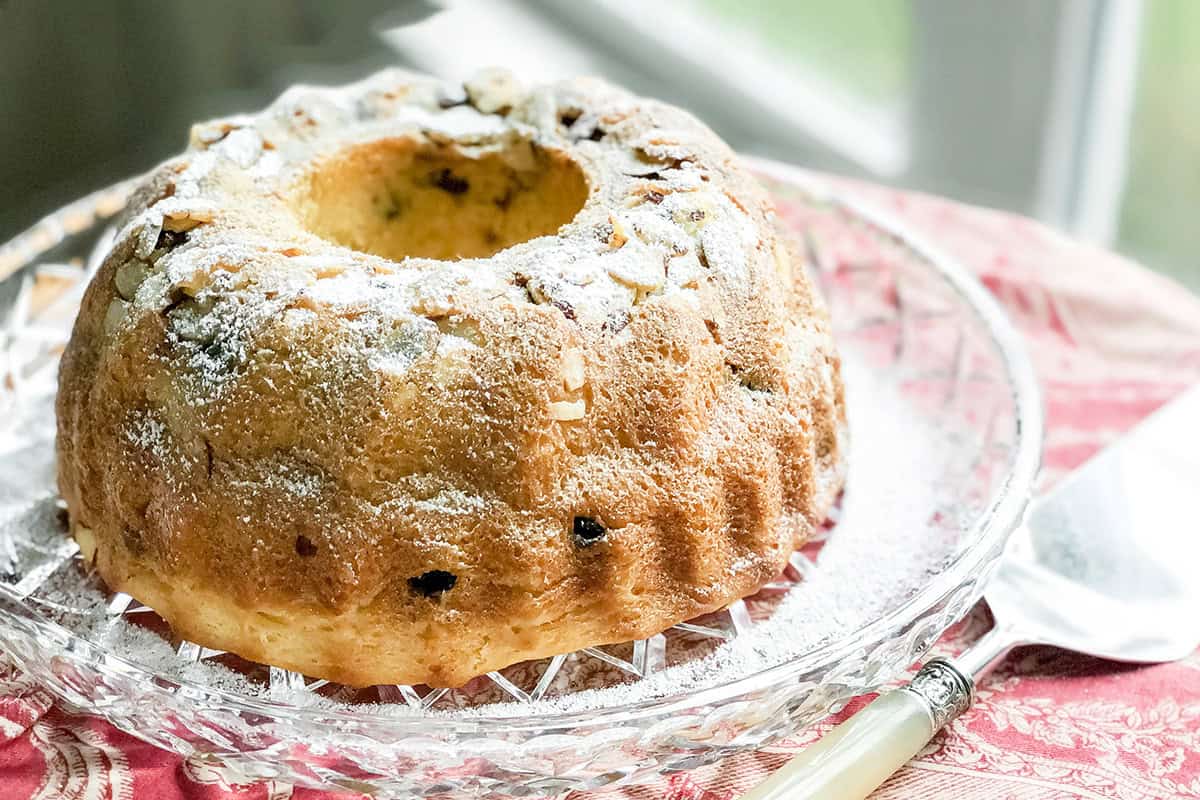A kugelhopf bundt cake dusted with powdered sugar sits on a glass platter next to a silver knife, placed on a red patterned tablecloth.