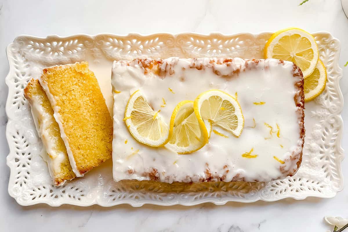 A loaf of lemon drizzle cake with white icing and lemon slices on top, served on a decorative white platter with two slices cut.