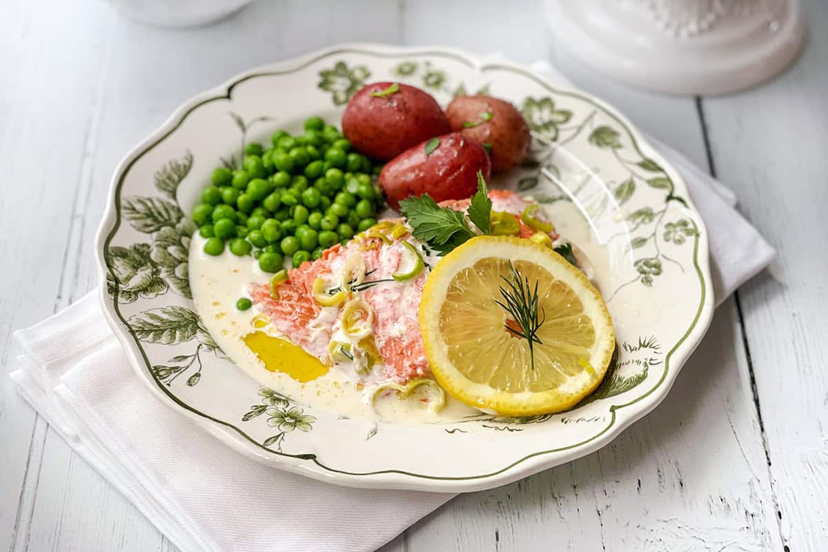 A plate with baked salmon topped with a lemon slice and dill, green peas, and red potatoes, served on a floral dish with a white napkin underneath.