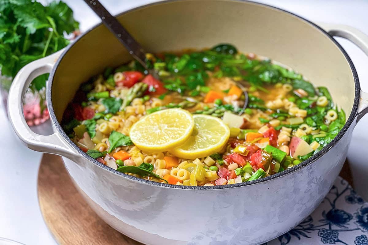 A pot of spring minestrone soup with ditalini pasta, spinach, tomatoes, and two lemon slices on top, with a ladle inside the pot.