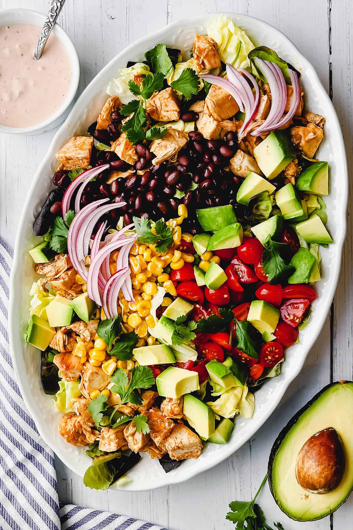 An oval platter with rows of lettuce, grilled chicken, black beans, corn, avocado, cherry tomatoes, red onion, and cilantro captures the essence of a fresh chicken taco salad. Served with a bowl of dressing and half an avocado.