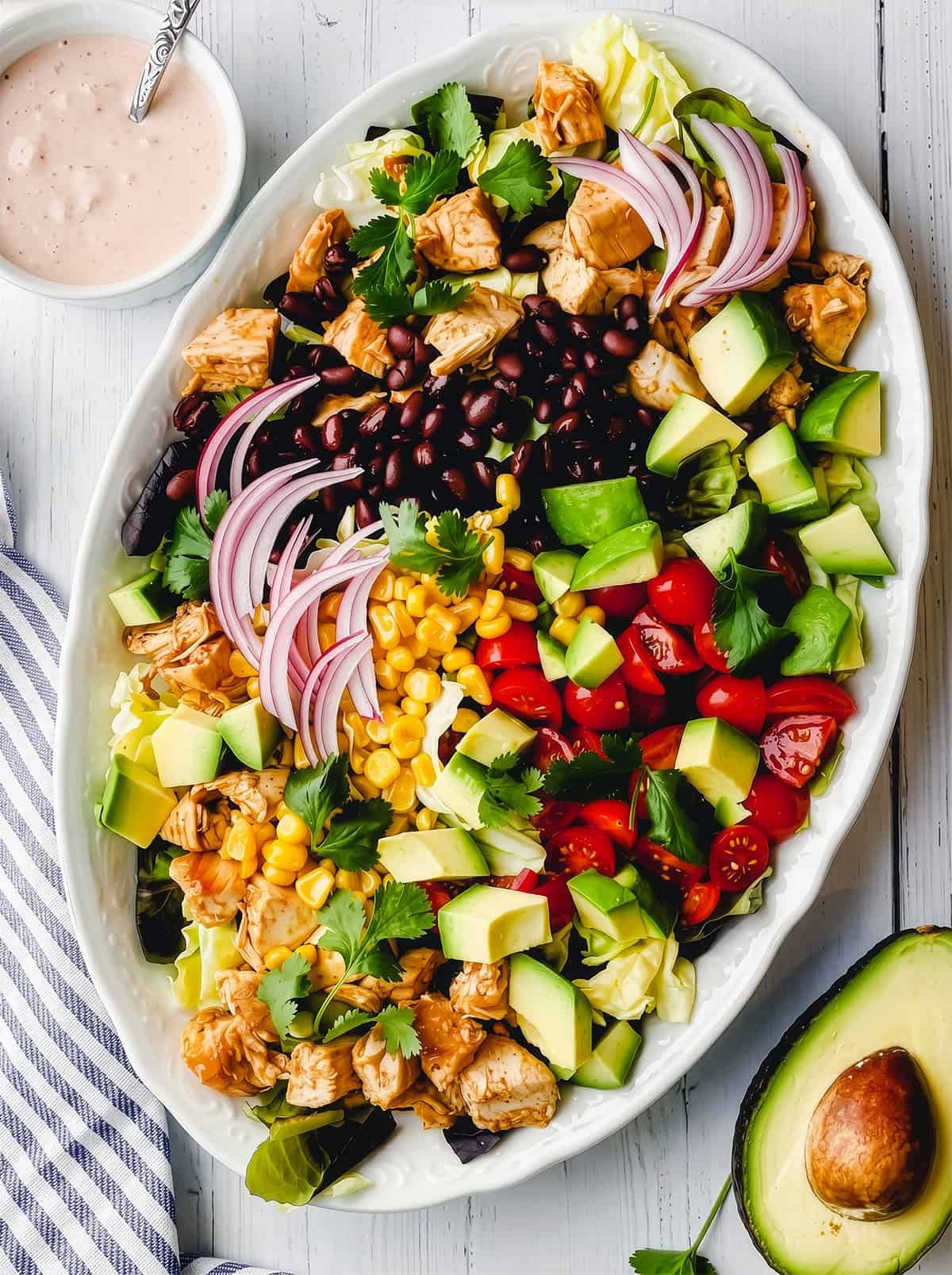 Oval platter with rows of chicken, black beans, corn, tomatoes, avocado, red onion, greens, and cilantro for an easy chicken taco salad recipe. Served with a small bowl of dressing and half an avocado on the side.