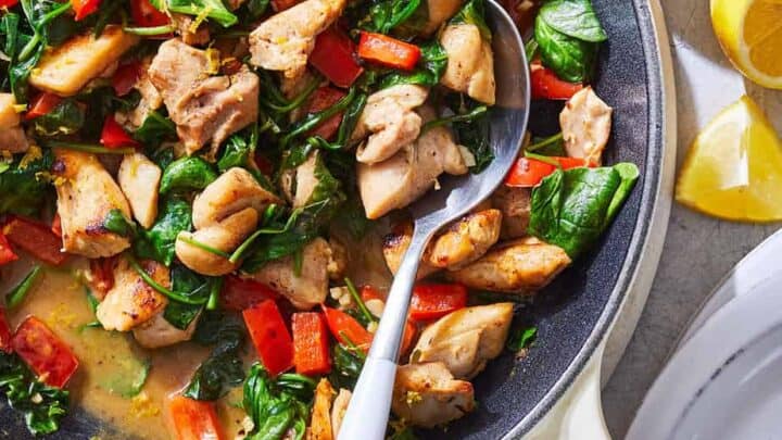 A skillet filled with cooked chicken pieces, red bell peppers, and spinach makes for a perfect Spring One Pot Dinner; lemon wedges and grated cheese are served on the side.