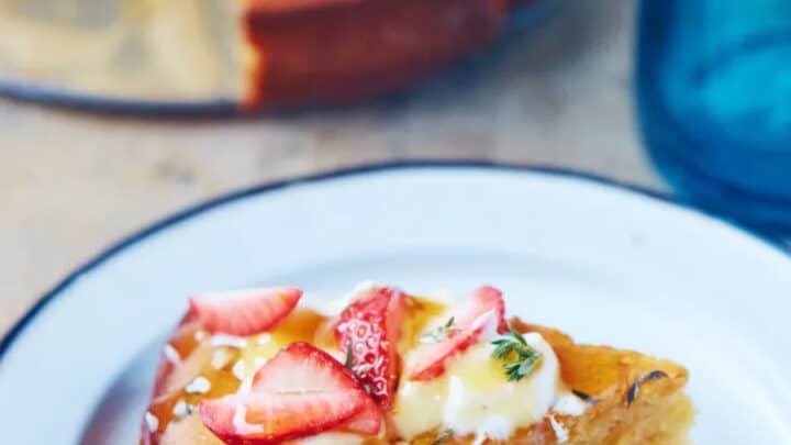 A slice of yellow cake topped with sliced strawberries, cream, and syrup sits on a white plate; the remaining cake is in the background—a delightful treat to follow your April Mediterranean Diet Dinners.