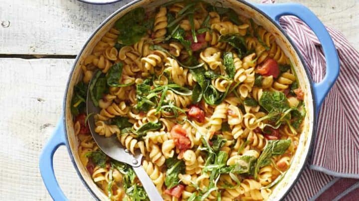 A blue pot filled with rotini pasta, tomatoes, and leafy greens sits on a white wooden table with a serving spoon&mdash;perfect for Spring One Pot Dinners; a plate of cheese and basil leaves are nearby.