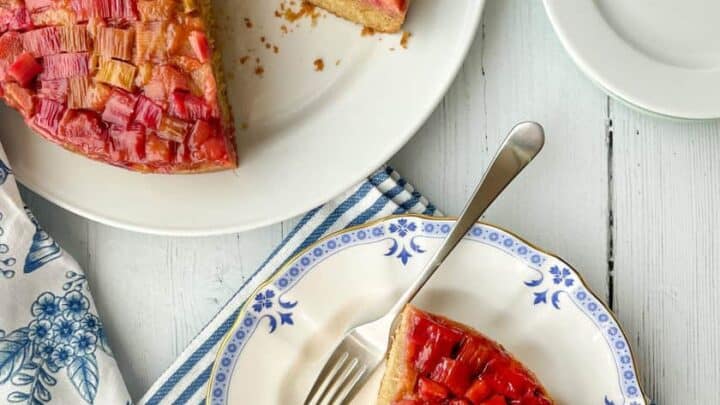 Top view of a slice of Rhubarb Upside Down Cake on a blue and white plate with the whole cake in the top left hand corner.