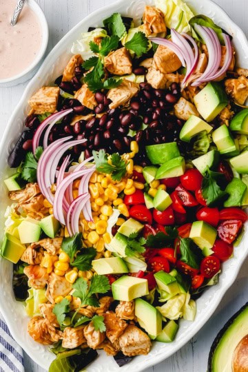 Oval platter with chopped lettuce, chicken, black beans, avocado, cherry tomatoes, corn, red onion, and cilantro—this chicken taco salad recipe is served with a creamy dressing on the side.