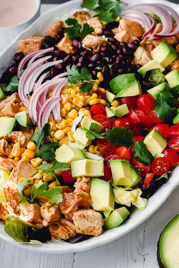 A chicken taco salad platter with diced chicken, avocado, cherry tomatoes, corn, black beans, red onion, lettuce, and cilantro, served with a creamy dressing on the side.