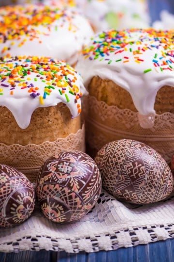 Iced Easter cakes, or kulich, with colorful sprinkles and intricately decorated Easter eggs are displayed on a lace cloth.
