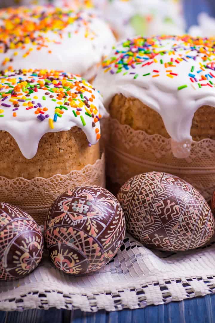 Iced Easter cakes, or kulich, with colorful sprinkles and intricately decorated Easter eggs are displayed on a lace cloth.
