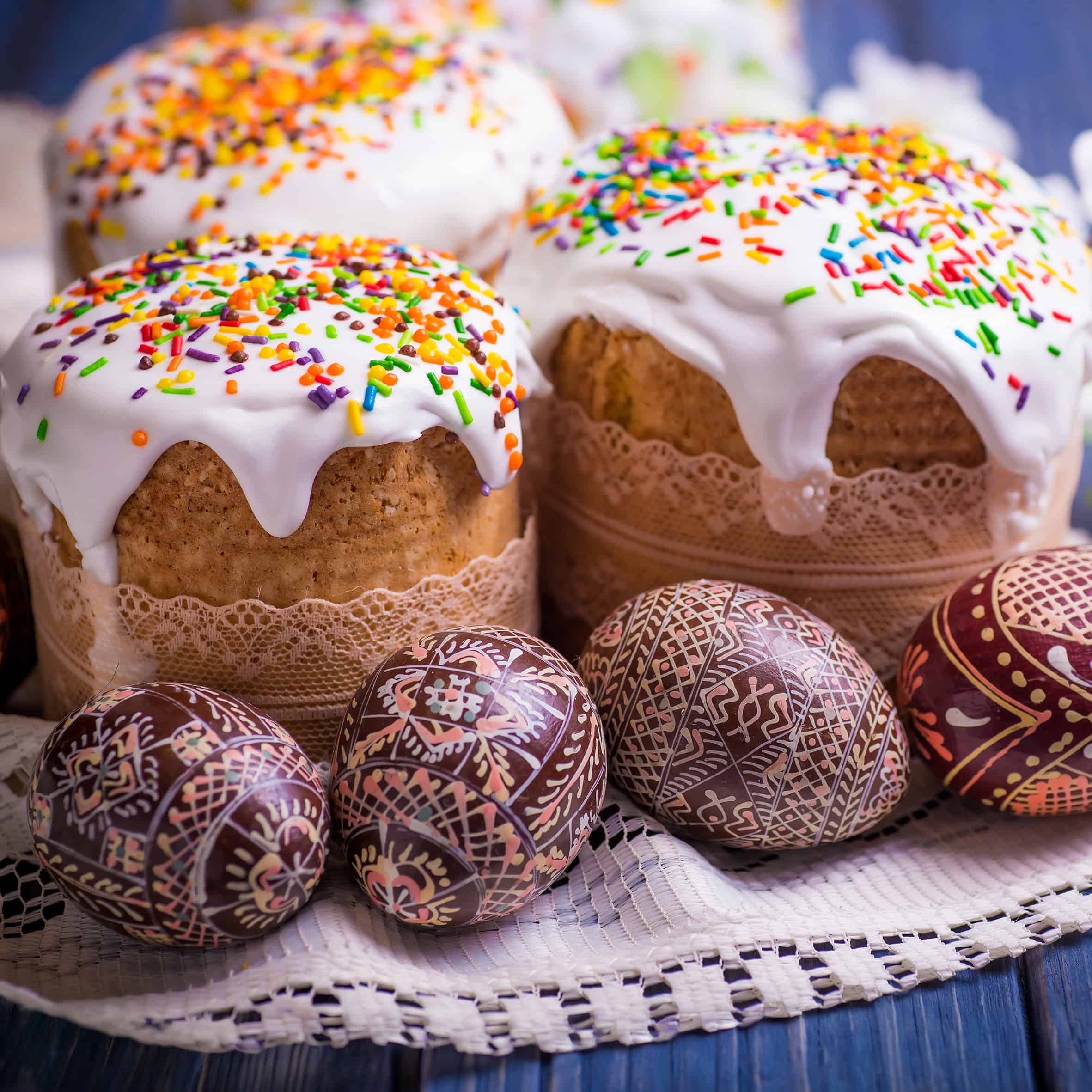Iced Easter cakes, or kulich, with colorful sprinkles and intricately decorated Easter eggs are displayed on a lace cloth.