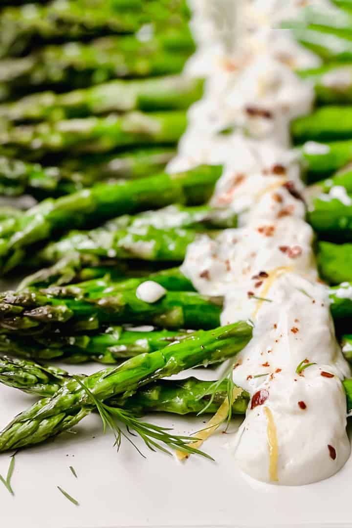 Close-up of roasted asparagus spears arranged on a platter, topped with a creamy white sauce and garnished with herbs and red pepper flakes for a flavorful roasted asparagus dish.