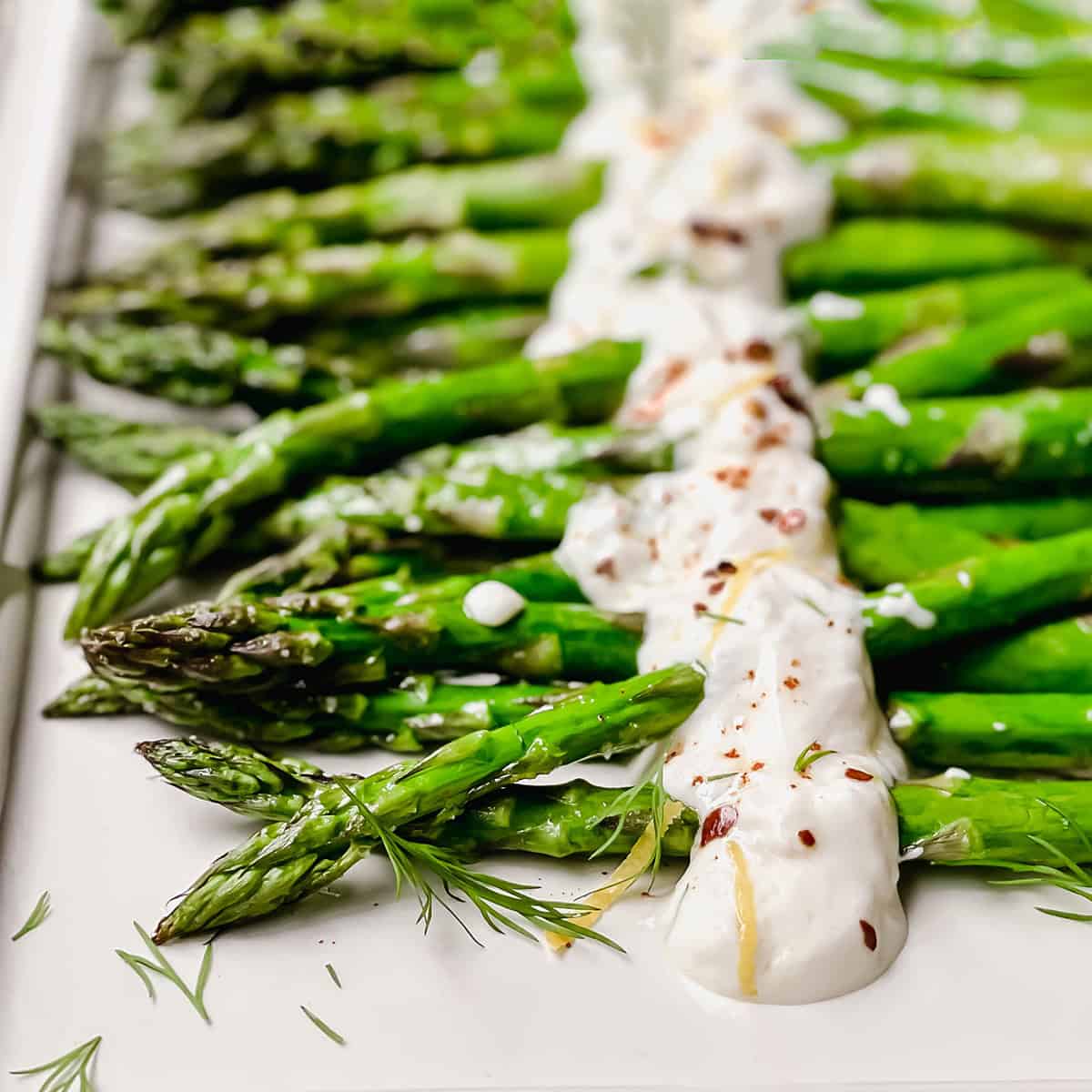 Close-up of roasted asparagus spears arranged on a platter, topped with a creamy white sauce and garnished with herbs and red pepper flakes for a flavorful roasted asparagus dish.