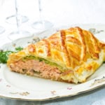 A slice of salmon wellington en croûte with a golden, flaky pastry crust rests on a decorative plate.