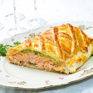 A slice of salmon wellington en croûte with a golden, flaky pastry crust rests on a decorative plate.