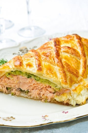 A slice of salmon wellington en croûte with a golden, flaky pastry crust rests on a decorative plate.
