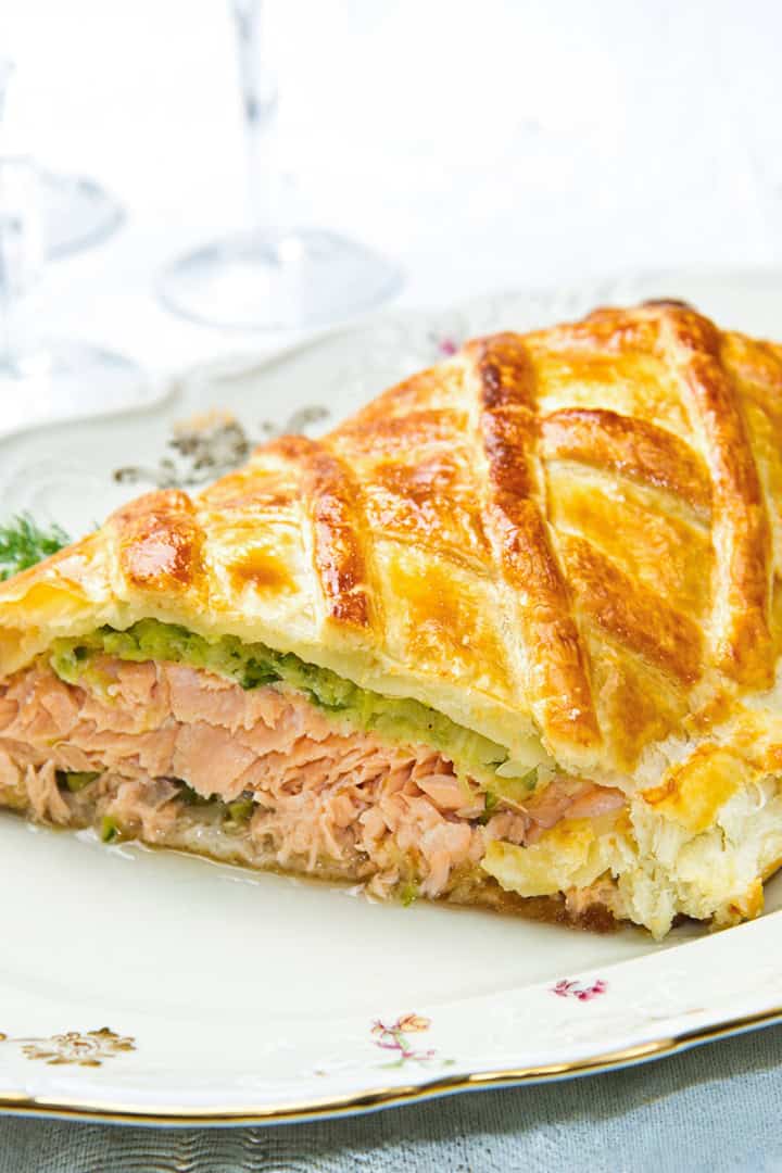 A slice of salmon wellington en croûte with a golden, flaky pastry crust rests on a decorative plate.