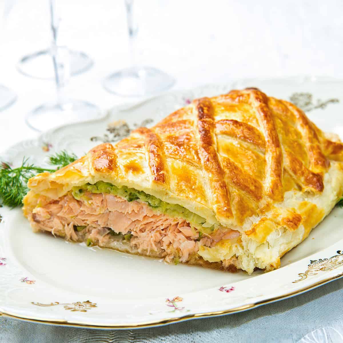 A slice of salmon wellington en croûte with a golden, flaky pastry crust rests on a decorative plate.