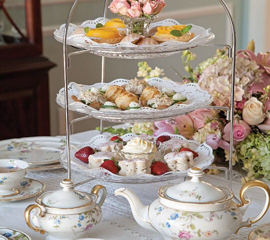 A three-tiered tray with assorted pastries and sweets is set on a lace tablecloth with floral china teapots, cups, and a bouquet of flowers-the perfect setting for a charming Mother's Day Tea.